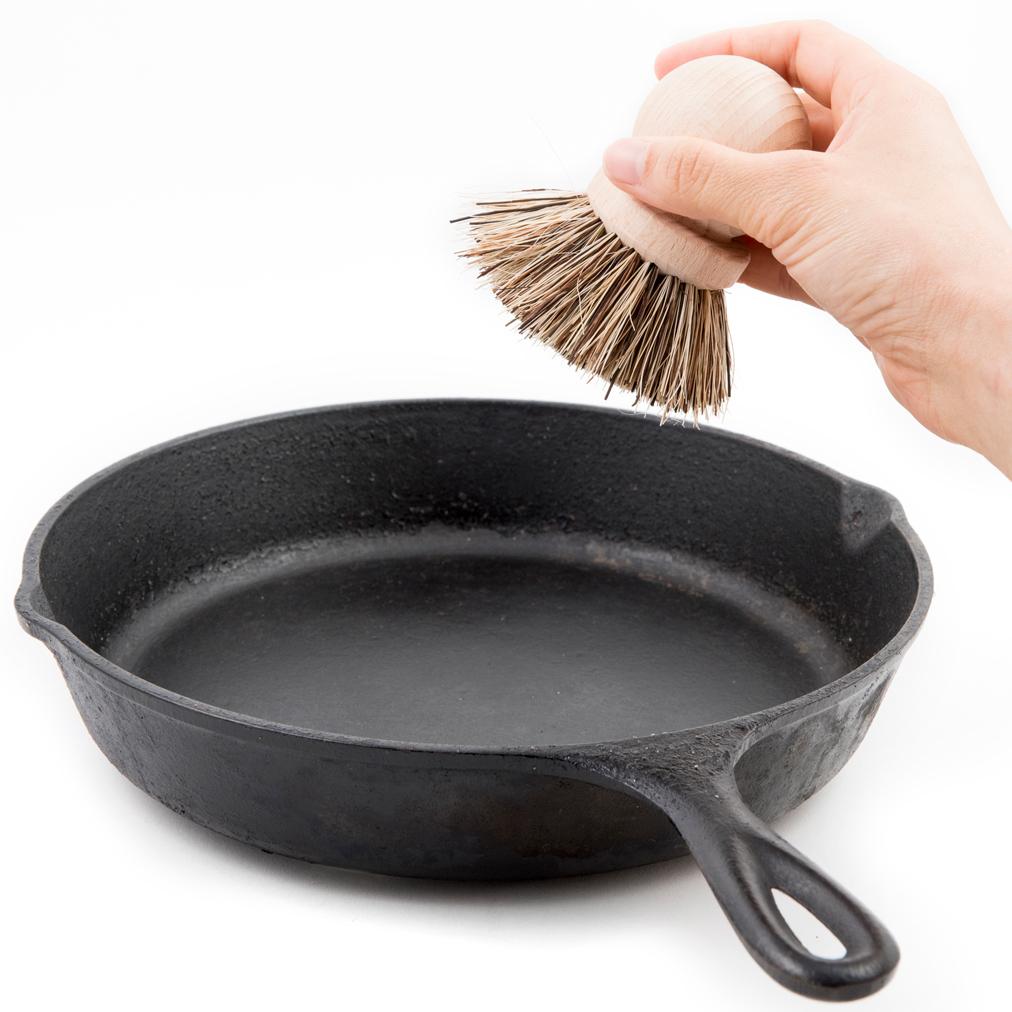 Hand holding wooden dish brush with natural bristles above a cast iron pan.