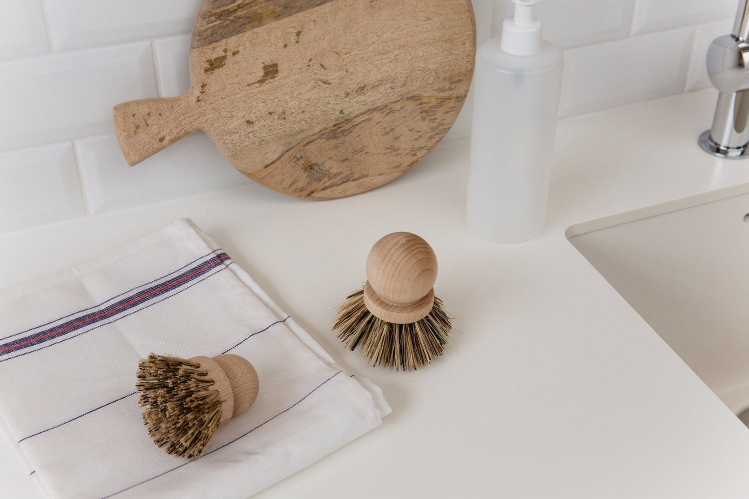 Two wooden dish brushes with natural bristles on counter next to sink and wooden cutting board.