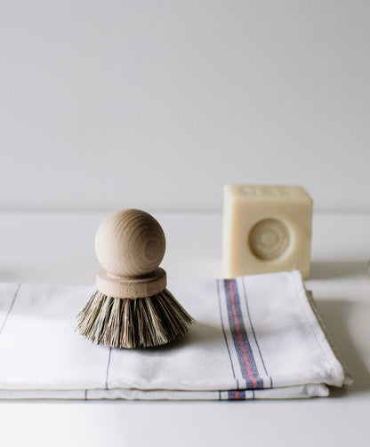 A wooden dish brush with natural bristles is displayed on a folded white dish towel with blue and red stripes. To the right, there&