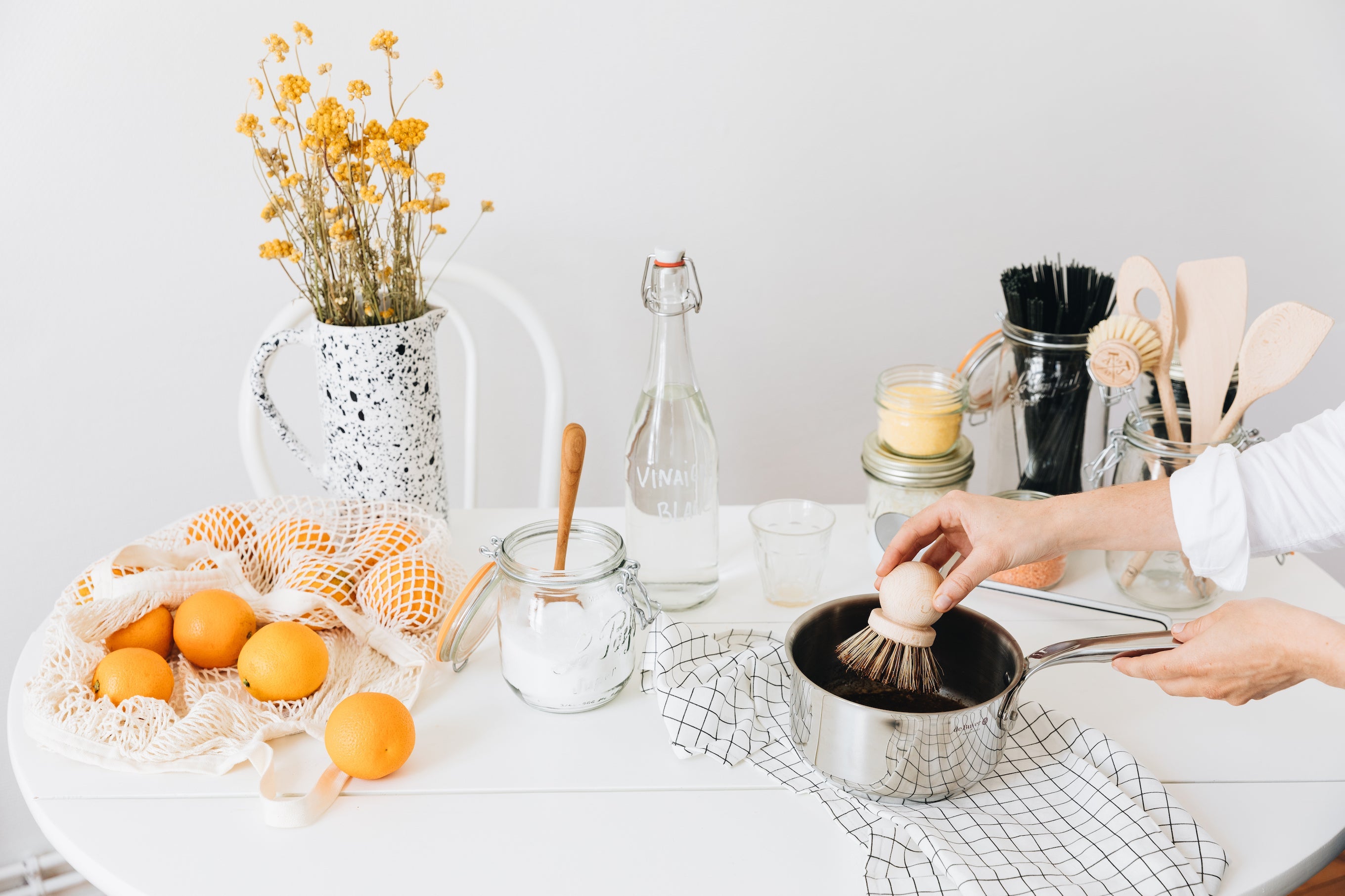 Bright table setting with fruits, flowers, kitchen utensils, and person scrubbing a dirty pot with a natural wooden dish scrub brush.