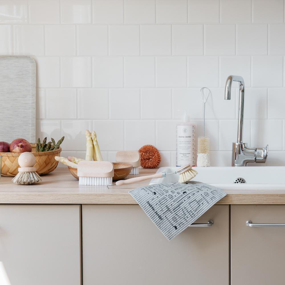 Kitchen counter covered with dish brushes, bottle brushes, scrub brushes, copper sponge, vegetables in bowls, dish soap, all next to sink.