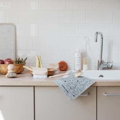 Kitchen counter covered with dish brushes, bottle brushes, scrub brushes, copper sponge, vegetables in bowls, dish soap, all next to sink.
