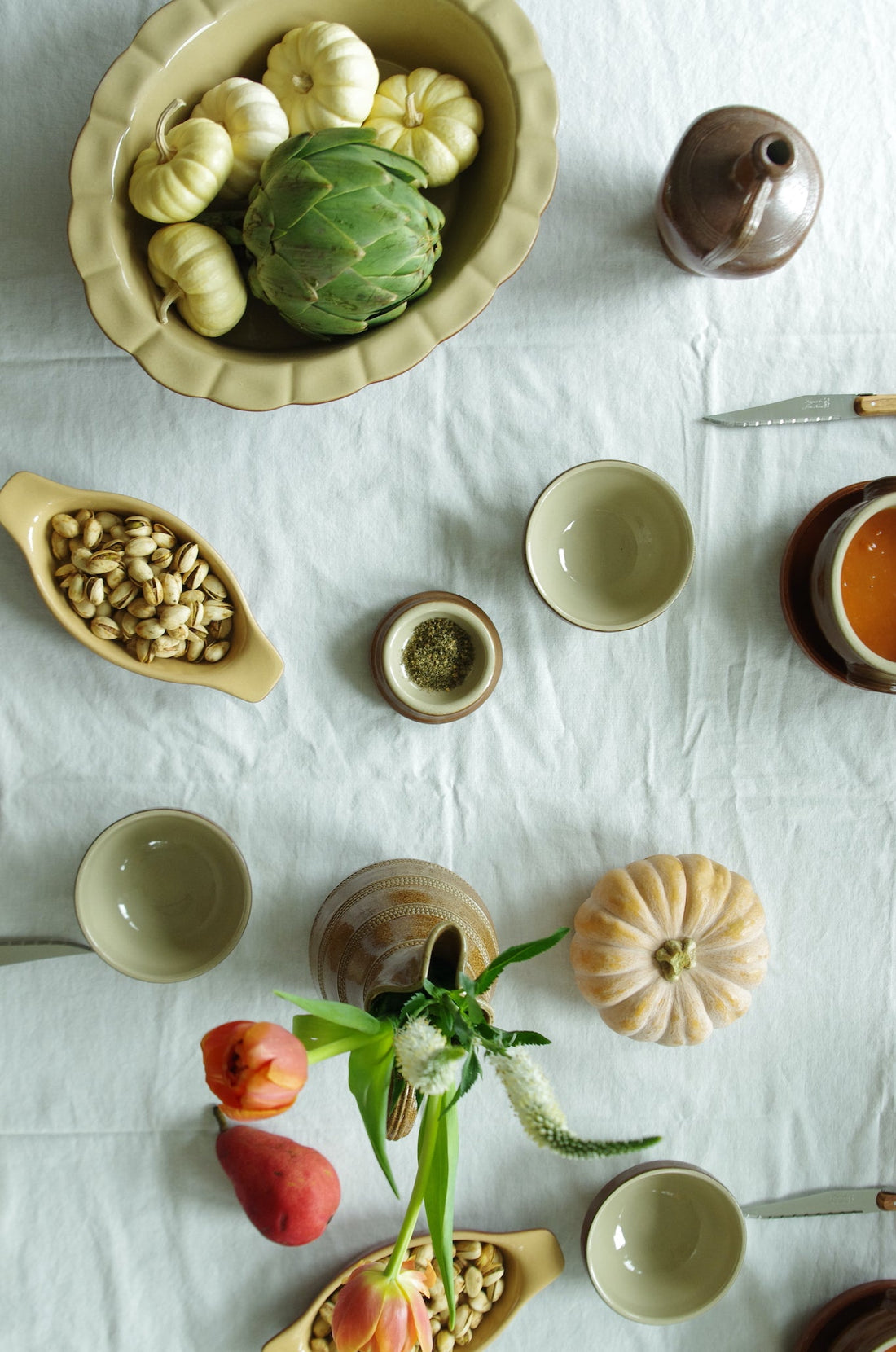 Tablescape photo taken of above. Table has a white natural tablecloth and displayed are French vintage pottery pieces by Poterie Renault including an olive oil jug or pitcher, serveware, bowls, and jugs used as vases holding a small bouquet. Pears and Laguiole French knives are also placed on the table as well as a pumpkin as decor.